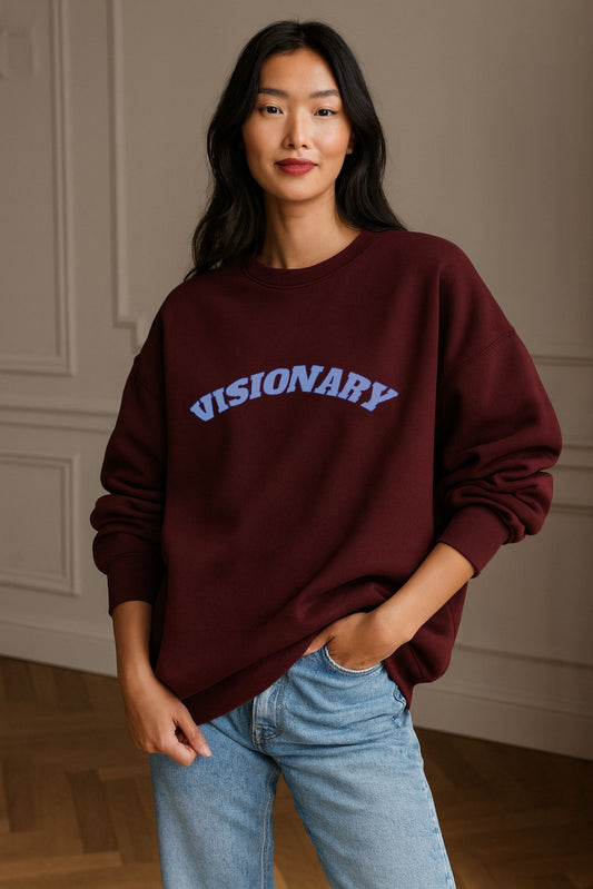 Model wearing FineAF VISIONARY sweatshirt in burgundy organic cotton fleece with soft blue text, minimalist studio light.