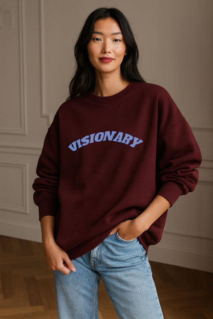 Model wearing FineAF VISIONARY sweatshirt in burgundy organic cotton fleece with soft blue text, minimalist studio light.