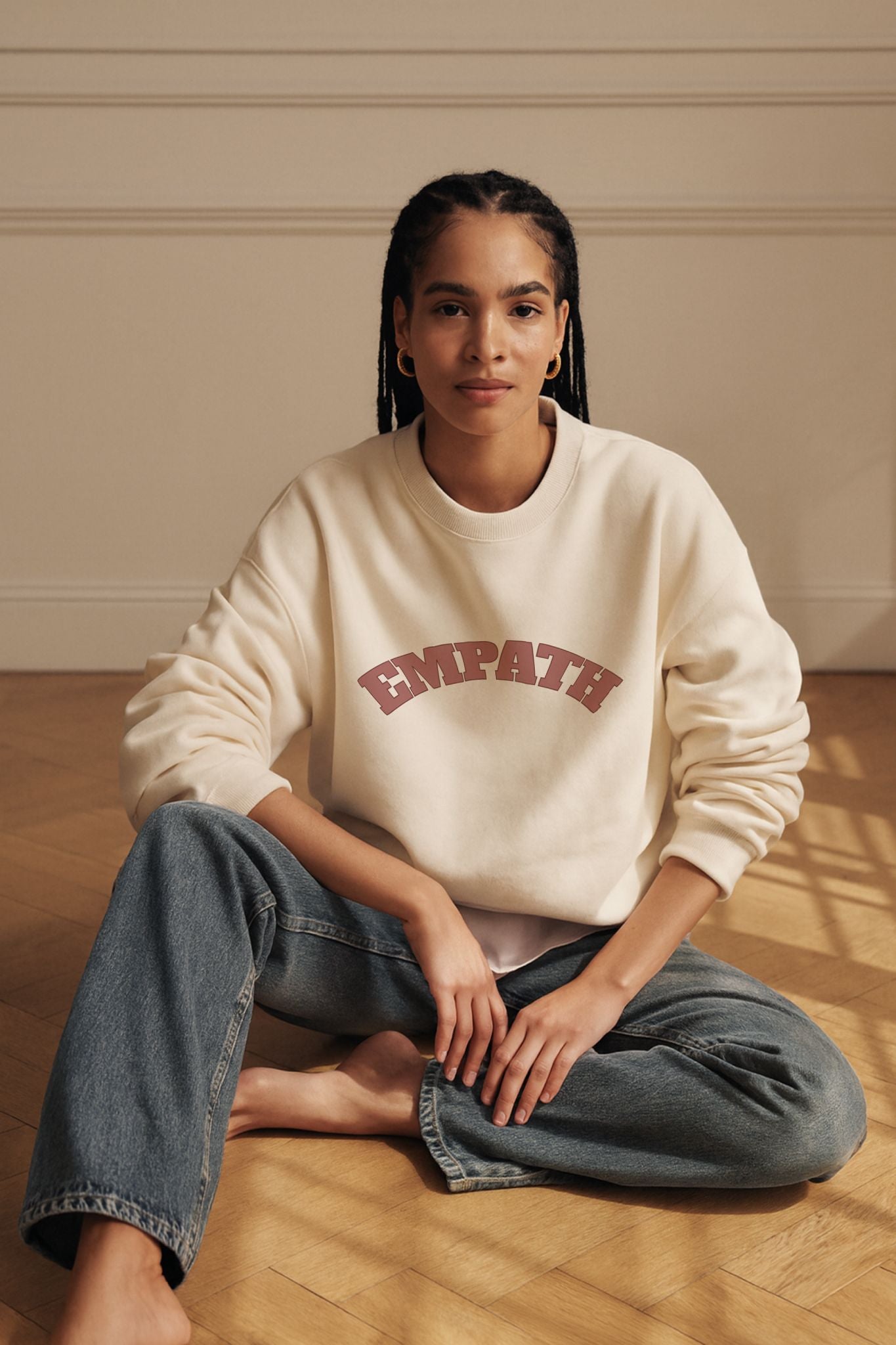 Woman seated on oak parquet floor wearing ivory EMPATH sweatshirt in organic cotton fleece with dusty rose text, minimalist studio lighting