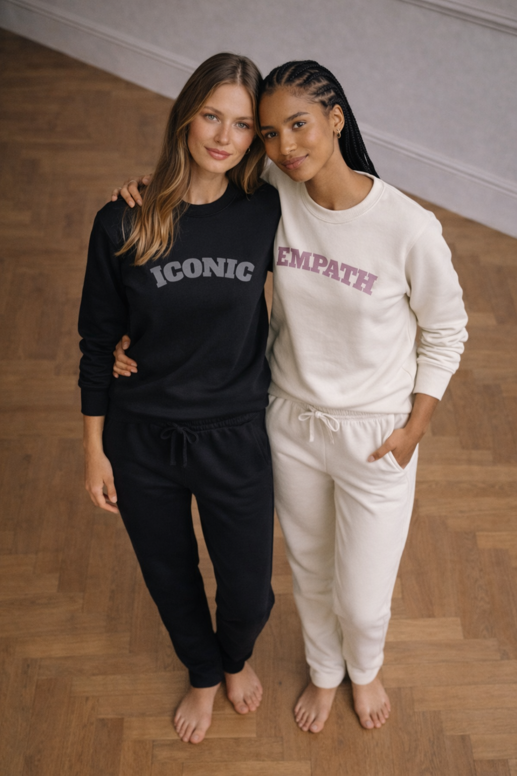 Two women wearing black and white sweatsuits with 'Iconic' and 'Empath' text on a wooden floor.