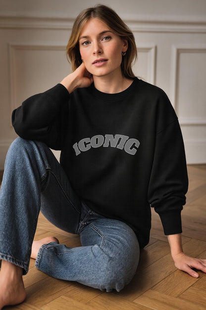 Black iconic crewneck sweatshirt in heavyweight organic cotton fleece, featuring a relaxed unisex fit, worn by a model seated on a wooden floor in daylight.